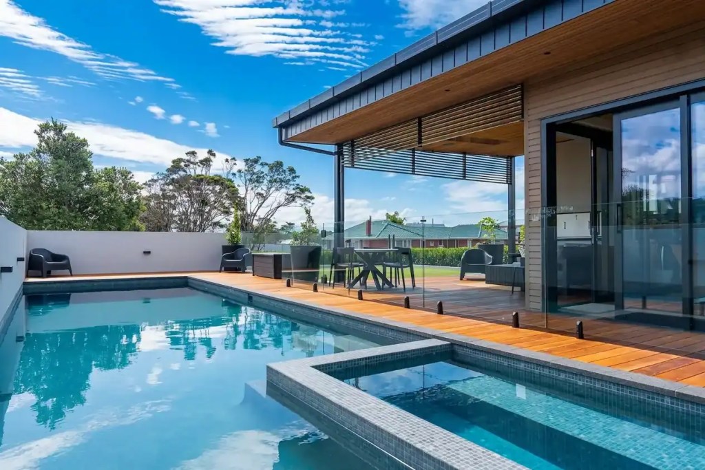 Seamless indoor-outdoor flow leading to a custom swimming pool and spa with glass fencing, timber decking, and a covered architectural entertaining area
